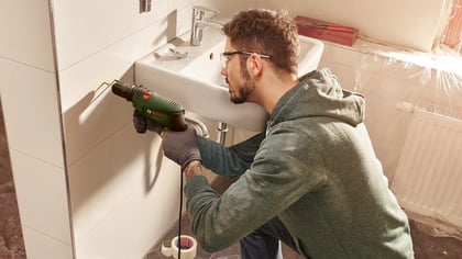 Un hombre usa un taladro Bosch verde para hacer un agujero en una pared de baño alicatada junto a un lavabo.