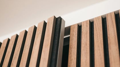 Primer plano de una pared de listones de madera con paneles acústicos. Los listones son de color marrón claro, el fondo es negro, y el conjunto está fijado a un techo blanco.