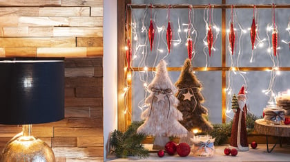 Weihnachtliche Fensterdekoration mit beleuchteter Lichterkette, roten Ornamenten, dekorativen Tannenbäumen, Holzscheiben, Kerzen und einer Santa Figur.