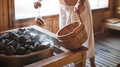 Person gießt Wasser mit einer Holzkelle auf heiße Saunasteine und erzeugt Dampf in einer Sauna.