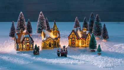 Festliche Weihnachtsdorf-Kulisse mit beleuchteten Häusern, Miniaturbäumen und Sängern im Kunstschnee.