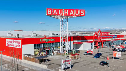 Vista aérea de una gran tienda Bauhaus de bricolaje y jardín roja y blanca con un letrero prominente, múltiples entradas y un concurrido estacionamiento.