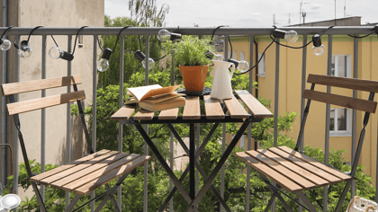 Balkontisch und zwei Stühle aus Holzlatten auf einem Balkon, dekoriert mit Lichterketten und Topfpflanzen, schaffen einen gemütlichen Außenbereich.