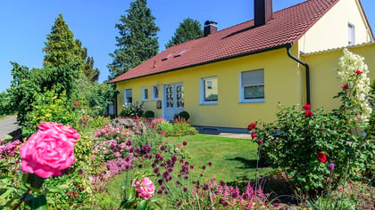 Ein hellgelbes Haus mit rotem Dach und buntem Garten, darunter rosa Rosen, neben einem gepflasterten Weg an einem sonnigen Tag.