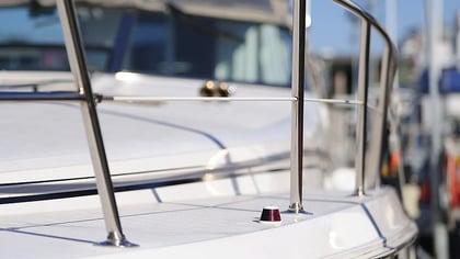 Detalle de una cubierta de barco blanca con barandillas cromadas y una pequeña luz de navegación roja, con un fondo de marina difuminado.
