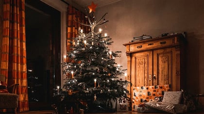 Ein beleuchteter Weihnachtsbaum, geschmückt mit weißen Lichtern, kerzenartigen Ornamenten und einem Stern, umgeben von Geschenken in einem schwach beleuchteten Wohnzimmer.