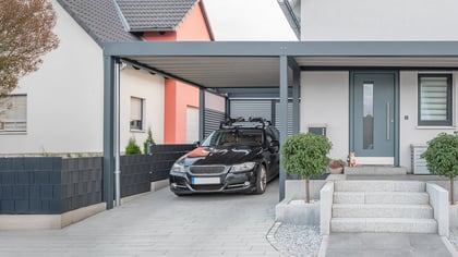 Modernes Haus mit dunklem Carport, das ein schwarzes Auto schützt. Helle Fassade, graue Tür und Steinstufen.