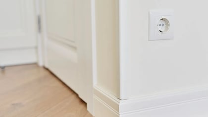 Enchufe eléctrico blanco en pared, junto a marco de puerta y rodapié blancos, sobre suelo de madera.
