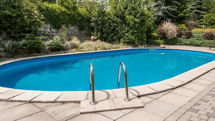 Ovaler blauer Swimmingpool mit hellen Pflastersteinen und üppigem Garten, zwei Liegestühle sichtbar.