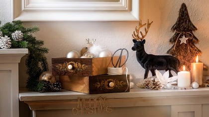 Weihnachtskamin mit immergrünen Girlanden, Tannenzapfen, Holzschatullen, Kerzen, einer Hirschfigur und einem 'Merry Christmas'-Schild dekoriert.