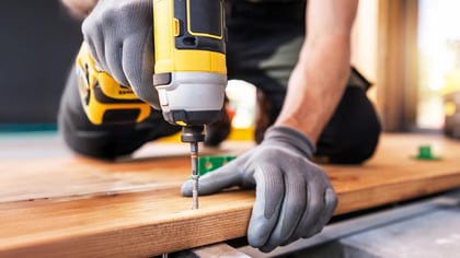 Ein Handwerker mit grauen Handschuhen, der eine gelbe Bohrmaschine benutzt, um eine Schraube in ein Holzbrett zu drehen.