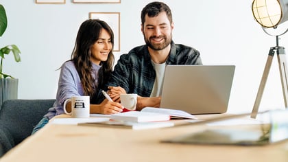 Zwei lächelnde Personen arbeiten gemeinsam an einem Laptop an einem Schreibtisch im modernen Heimbüro.
