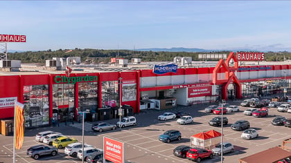 Vista aérea de una gran tienda de mejoras para el hogar Bauhaus roja con un estacionamiento lleno bajo un cielo despejado.