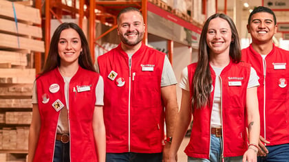 Cuatro empleados sonrientes de ferretería con chalecos rojos en un pasillo con madera apilada.