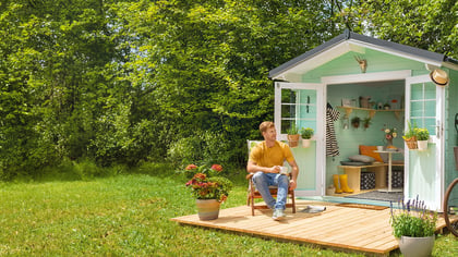 Ein hellgrünes Gartenhaus mit geöffneten Türen, davor ein Mann im Sessel und eine Frau mit Fahrrad. Ideal für Gartenliebhaber.