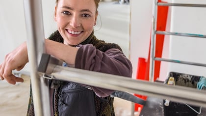 Mujer sonriente apoyada en un andamio, rodeada de herramientas de renovación.