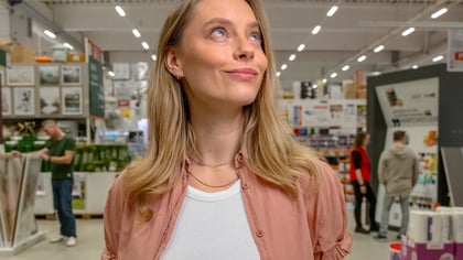 Joven rubia con camisa rosa, mirando pensativamente hacia arriba en un pasillo de tienda iluminado.