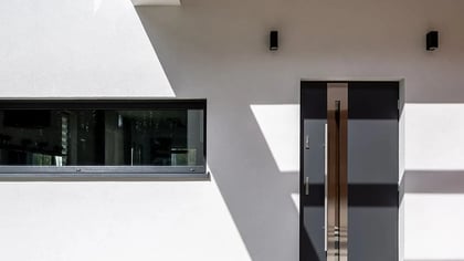 Moderna puerta principal gris oscuro con manija larga plateada junto a una ventana horizontal en una fachada blanca.