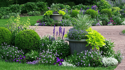 Ein gewundener Ziegelweg in einem üppigen Garten, umgeben von grünen Pflanzen, bunten Blumen und großen Dekotöpfen.