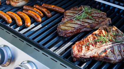 Parrilla de gas de acero inoxidable con rejillas negras, cocinando filetes y salchichas. Los mandos son visibles.