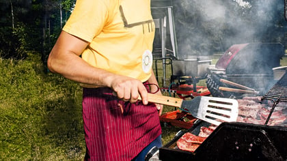 Ein Mann, der mit einem Pfannenwender Fleisch auf einem Grill wendet, wobei der Fokus auf dem Zubereitungsvorgang und dem Grillequipment liegt.