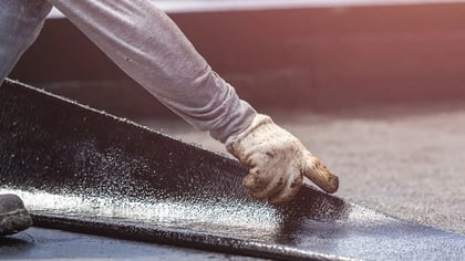 Trabajador instalando membrana asfáltica negra en techo plano, guiando el material con mano enguantada.
