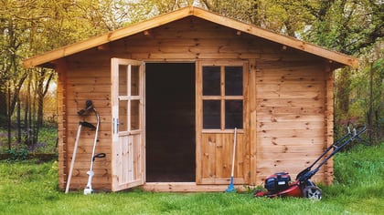 Caseta de jardín de madera con puerta abierta, rodeada de herramientas de jardinería y exuberante vegetación. Ideal para guardar todo.