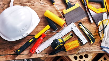 Bauwerkzeuge auf einem Holztisch, darunter ein Hammer, Schraubendreher, Maßband, Handschuhe und ein Schutzhelm.