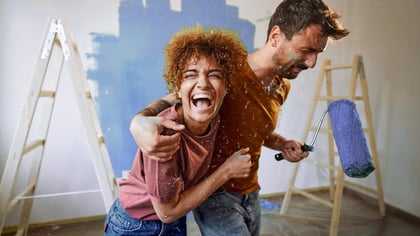 Una pareja riendo, cubierta de pintura azul, sosteniendo un rodillo, pintando una habitación.