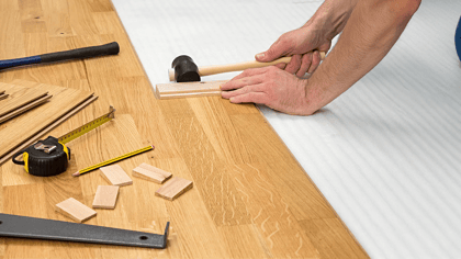 Bild von Laminatverlegungswerkzeugen und einer Person, die Laminat verlegt, mit Hammer, Bleistift, Maßband und Verlegeklötzen.