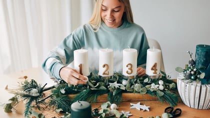 Eine Frau dekoriert einen Adventskranz mit vier nummerierten weißen Kerzen und einer separaten grünen Kerzenanordnung, umgeben von Grün und Ornamenten.