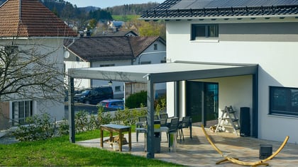 Pérgola gris moderna adosada a casa blanca, cubre una terraza con comedor.