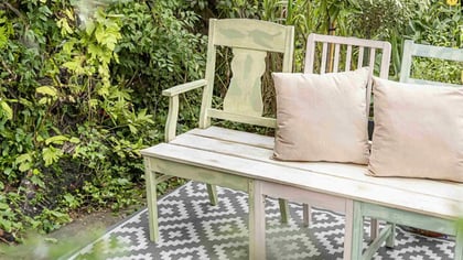 Gartenbank mit zwei Kissen und hellgrünem Anstrich, umgeben von üppigem Grün.