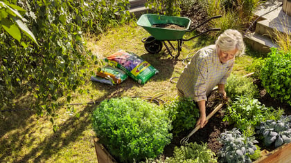 Mujer haciendo jardinería, usando una carretilla verde y sacos de tierra Gardigo en un huerto elevado.