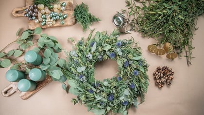 Materialien zum Basteln eines festlichen Kranzes: ein grüner Kranz, Eukalyptus, grüne Kerzen, Ornamente und Tannenzapfen.