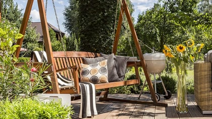Un acogedor patio exterior con un columpio de madera, un sofá de mimbre y una parrilla, situado en una cubierta de madera rodeada de plantas.