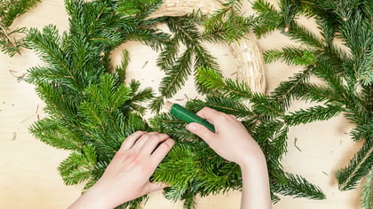 Die Hände einer Person, die einen Weihnachtskranz aus Tannenzweigen mit grünem Blumendraht bastelt, auf einem Holztisch.