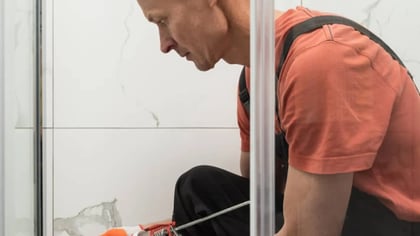 Una persona aplica sellador al borde de una mampara de ducha de cristal con una pistola de calafateo, sellándola contra azulejos blancos.