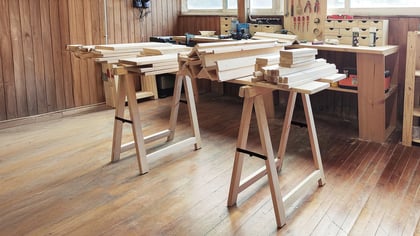 Zwei helle Holzböcke tragen Stapel unbehandelter Holzbretter in einer Werkstatt.