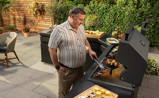 Mann grillt Essen auf einer schwarzen Außenküche auf einer Terrasse. Eine Frau deckt einen Tisch für Mahlzeiten im Freien.