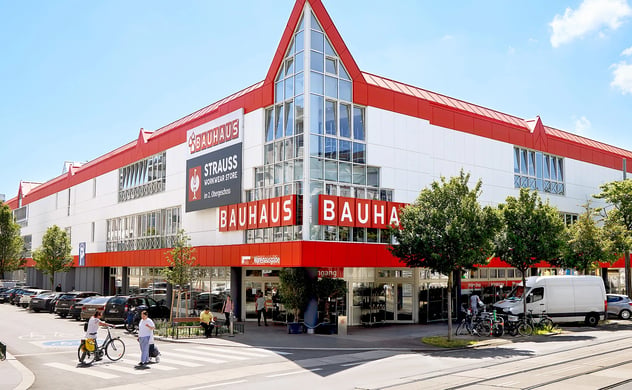 Außenansicht eines großen, rot-weißen BAUHAUS Fachcentrums und Straße mit Menschen, Autos, Fahrrädern und Straßenbahnschienen an einem sonnigen Tag.