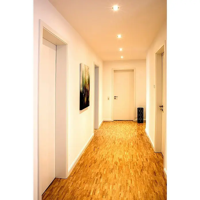 Müller-Licht Tint LED-Einbauleuchten-Set - 19 W, Weiß, 3 Stk., RGBW - Helle Fluransicht mit Parkettboden, weißen Wänden und Türen sowie modernen Deckenleuchten, die eine einladende Atmosphäre schaffen.