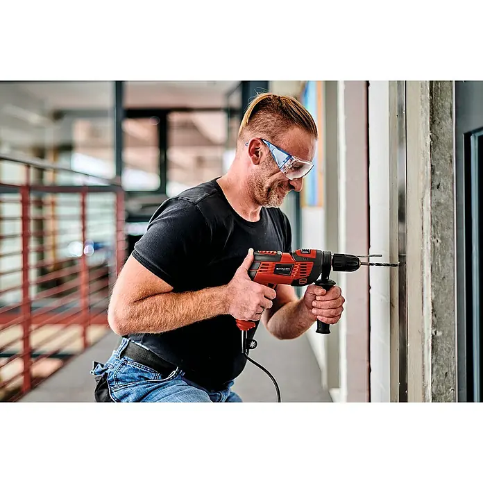 Einhell Schlagbohrmaschine TC-ID 720/1 E Kit - 720 W, Schnellspannbohrfutter - Ein Mann mit Schutzbrille bohrt mit einer Einhell Schlagbohrmaschine ein Loch in eine Wand. Das rote Werkzeug ist gut sichtbar.