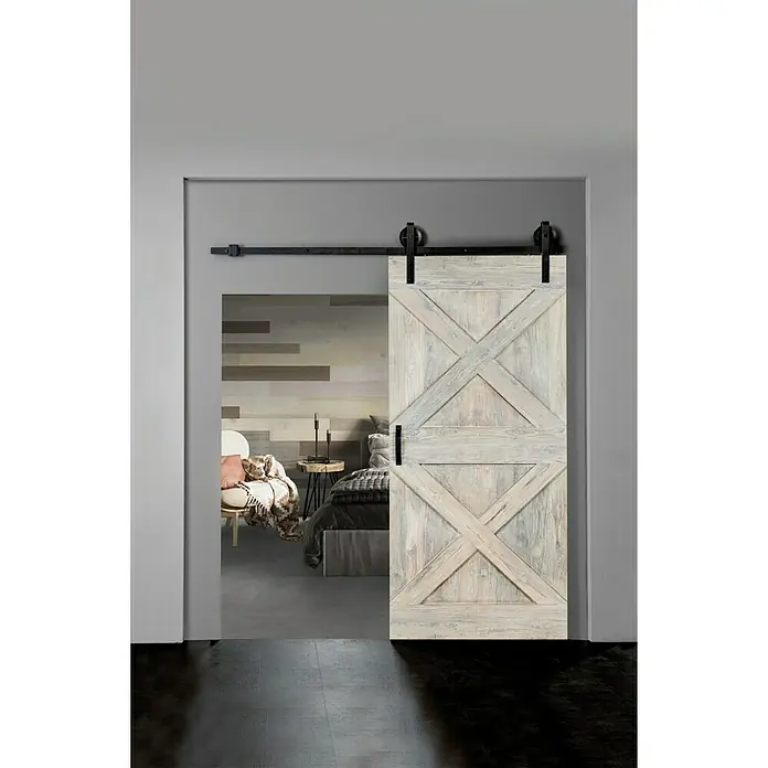 Indo Holzschiebetür Barn Door Colored - 100 x 215 cm, Teak, White washed - Eine helle Holztür im Scheunenstil, die teilweise einen Einblick in ein modernes Schlafzimmer mit einem eleganten Sessel und einem Bett bietet.