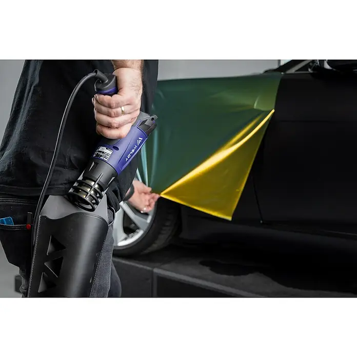 Weldy Heißluftgebläse HG 330-B Car-Wrap-Kit - 1.600 W, Temperaturbereich: 350 °C - 550 °C - Nahaufnahme eines Mannes, der mit einer WELDY Heißluftpistole eine gelbe Folie auf einem Auto anbringt.