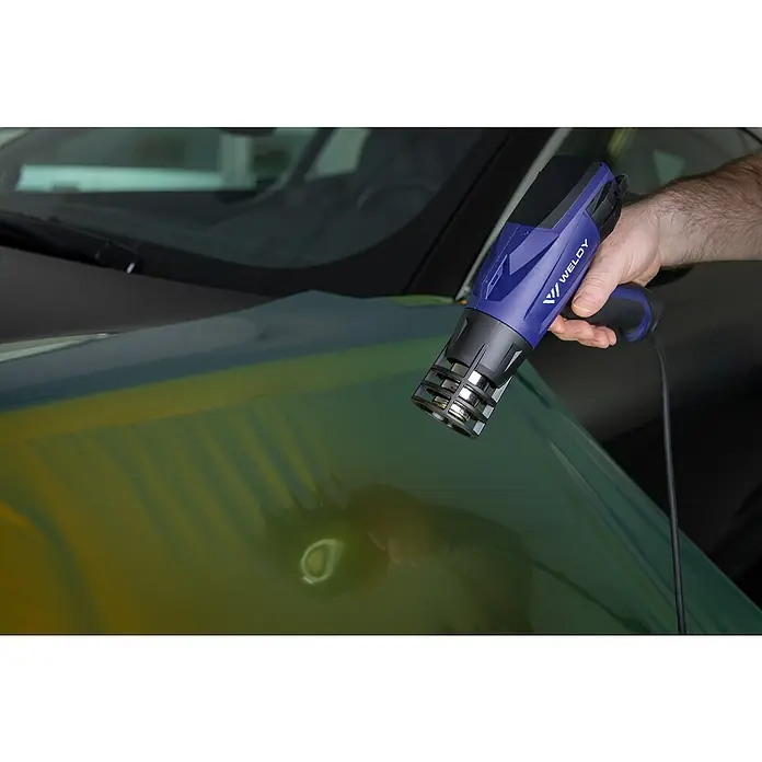 Weldy Heißluftgebläse HG 330-B Car-Wrap-Kit - 1.600 W, Temperaturbereich: 350 °C - 550 °C - Nahaufnahme einer blauen Weldy Heißluftpistole, die über eine mit grüner Folie umhüllte Motorhaube eines Autos gehalten wird.