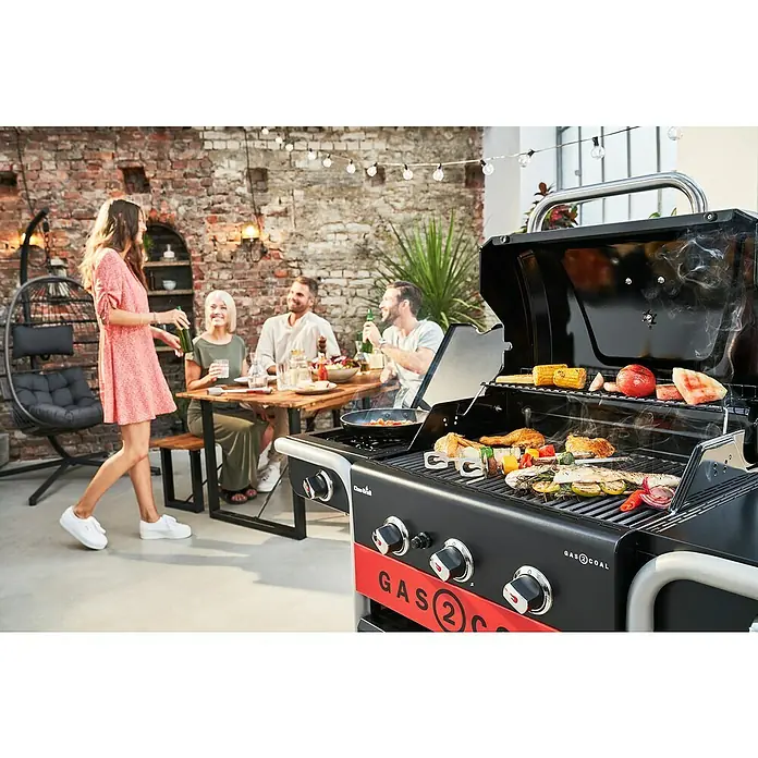Char-Broil Hybrid-Grill Gas2Coal 2.0 330Grillfläche (B x T): 61,5 x 44,1 cm, 10,1 kW, Anzahl Brenner: 3 - Der Gas2Coal Grill mit verschiedenem Grillgut wie Hähnchen, Gemüse und Maiskolben, während sich eine Gruppe von Menschen im Hintergrund entspannt.