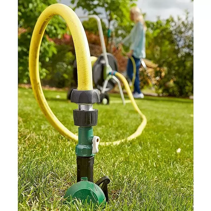 Wassersteckdose - 1″ - Nahaufnahme eines gelben Gartenschlauchs, der an einem grün-schwarzen Rasensprenger in einem Garten angeschlossen ist.