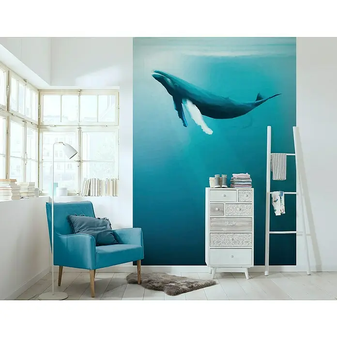 Komar Fototapete Artsy Humpback Whale - B x H: 200 x 280 cm, Vlies - Das blaue Sessel-Set mit einem Kissen, das vor einem weißen Fenster und einer Wand mit Walbild steht, schafft eine ruhige, maritime Atmosphäre.