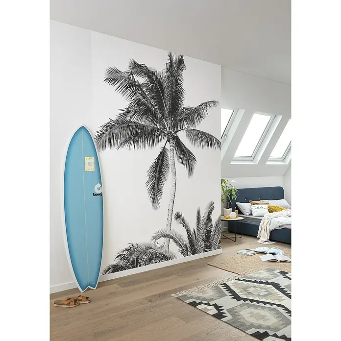 Komar Fototapete Retro PalmB x H: 200 x 280 cm, Vlies - Ein hellblaues Surfbrett lehnt an einer Wand mit einem Palmenmotiv in einem hellen, luftigen Schlafzimmer mit Holzböden.